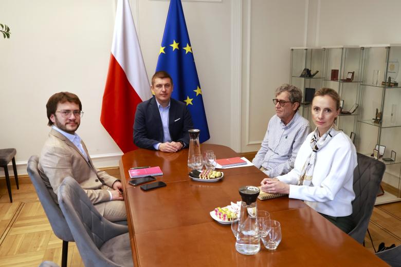 Cztery osoby pozują do wspólnego zdjęcia siedząc przy stole na tle flag Polski i UE