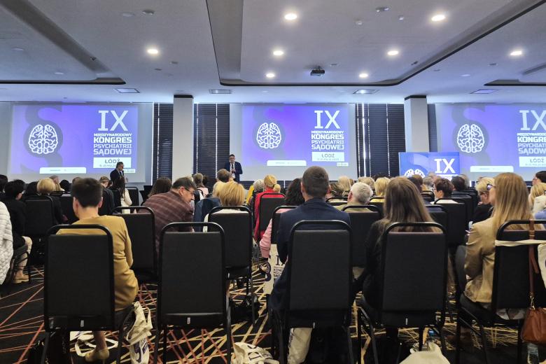 Kilkadziesiąt osób na dużej sali konferencyjnej siedzi na krzesłach i słucha wystąpienia osoby mówiącej z podwyższenia, w tle na trzech wyświetlaczach grafika z napisem "IX Międzynarodowy Kongres Psychiatrii Sądowej - Łódź 9-10 maja 2025