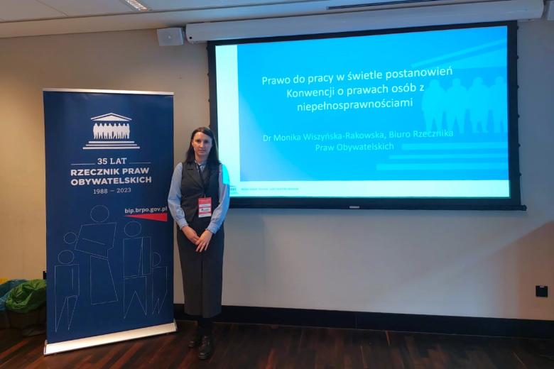 Osoba pozuje do zdjęcia obok rollupa z logotypem Rzecznika Praw Obywatelskich i wyświetlacza z tytułem wystąpienia "Prawo do pracy w świetle postanowień Konwencji o prawach osób z niepełnosprawnościami"