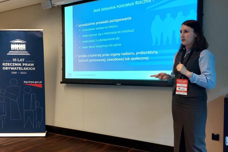 Osoba mówi do mikrofonu na tle wyświetlacza z prezentacją