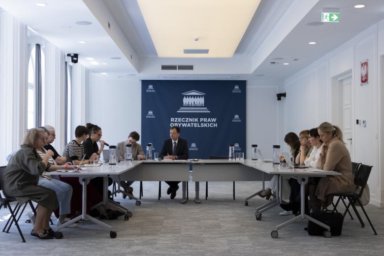 Kilkanaście osób siedzi przy stołach ustawionych w podkowę, za stołami przy ścianie rząd flag Polski i UE