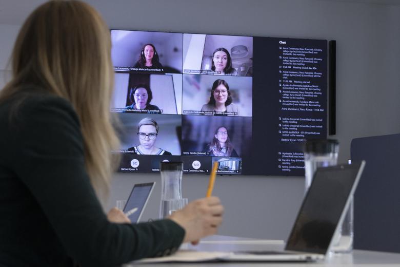 Osoba siedząca za stołem spogląda w duży wyświetlacz z widocznymi uczestnikami spotkania łączącymi się za pomocą programu komputerowego do komunikowania się na odległość
