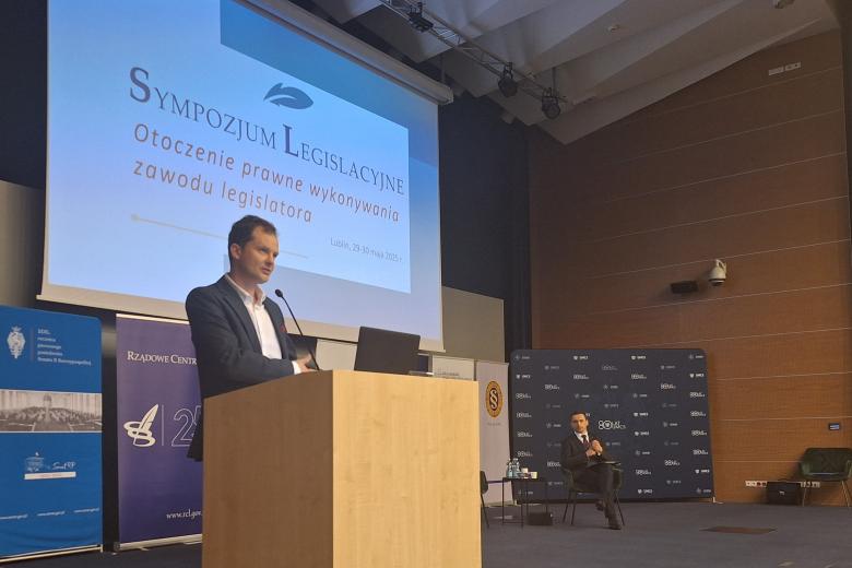 Adam Krzywoń wypowiada się, stojąc przy mównicy w audytorium. Za nim znajduje się ekran projektora wyświetlający nazwę wydarzenia: "Sympozjum Legislacyjne - Otoczenie prawne wykonywania zawodu legislatora"
