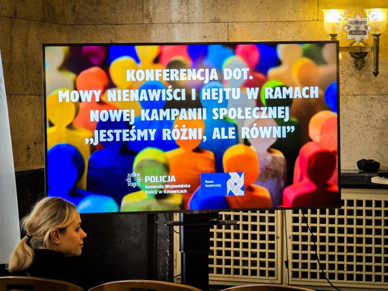 Ekran multimedialny, na którym wyświetla się slajd z napisem: Konferencja dot. mowy nienawiści i hejtu w ramach nowej kampanii społecznej "Jesteśmy różni, ale równi" i kolorowymi sylwetkami osób