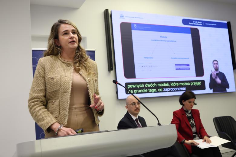 Beata Baran-Wesołowska wypowiada się przy mównicy. W tle widać innych uczestników panelu oraz ekran wyświetlający prezentację.