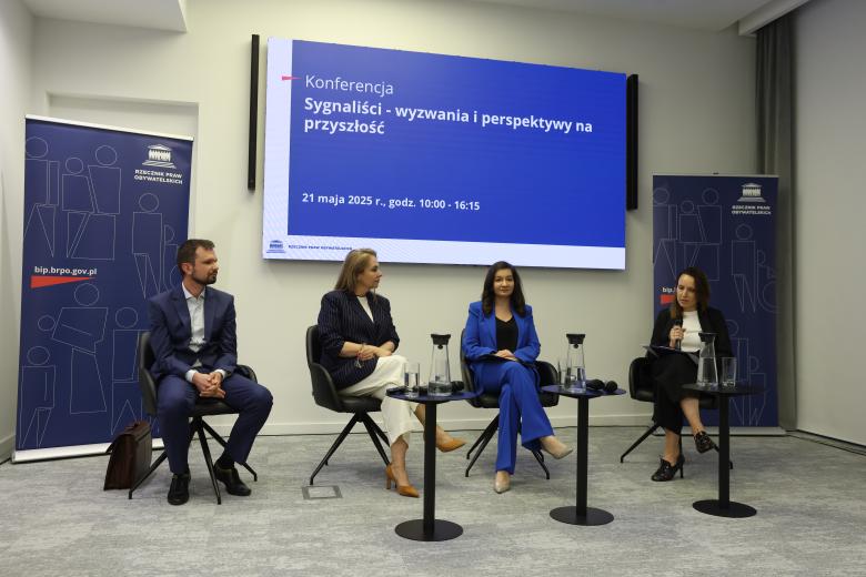 Czworo uczestników panelu siedzi na krzesłach ustawionych w szeregu. Za nimi znajduje się ekran wyświetlający planszę z napisem "Konferencja - Sygnaliści - wyzwania i perspektywy na przyszłość"