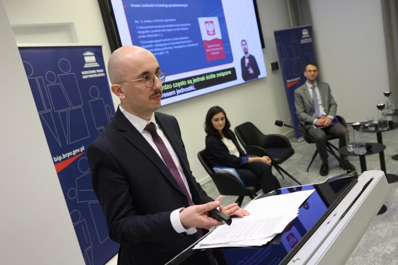 Marcin Malecko wypowiada się, stojąc przy mównicy. Obok niego siedzi dwoje innych uczestników panelu. W tle znajduje się ekran wyświetlający prezentację