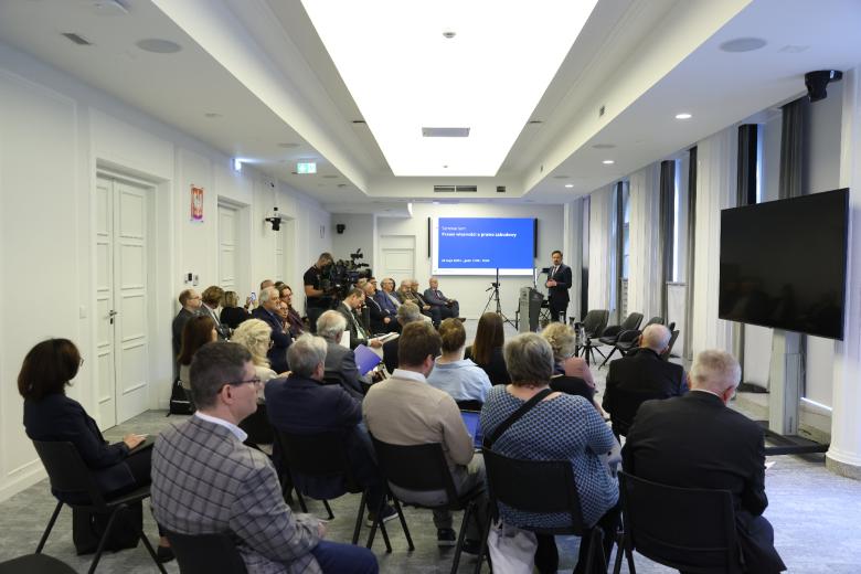 Widok ogólny sali konferencyjnej, w której uczestnicy seminarium siedzą na krzesłach ustawionych w rzędach. Przed widownią przy mównicy stoi i wypowiada się Marcin Wiącek