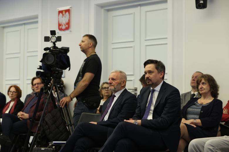 Uczestnicy seminarium siedzą na krzesłach i słuchają wypowiedzi uczestników sesji plenarnej. Między nimi stoi kamera telewizyjna i jej operator