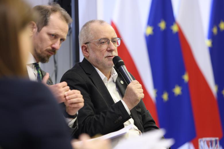 Piotr Tuleja wypowiada się do mikrofonu, siedząc obok innych uczestników sesji plenarnej