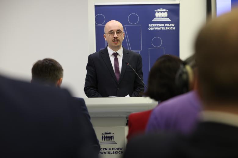 Marcin Malecko wypowiada się stojąc przy mównicy. Na pierwszym planie widać sylwetki siedzących na widowni osób