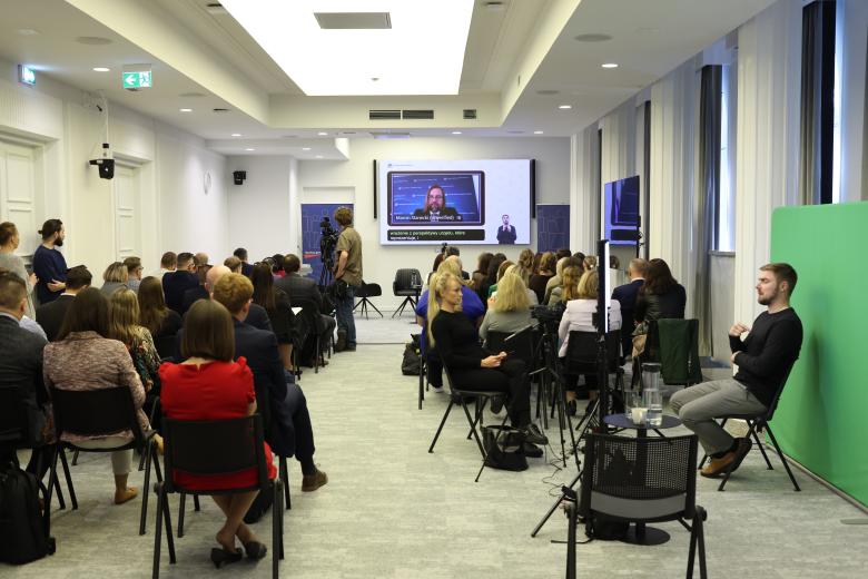 Sala konferencyjna wypełniona siedzącymi ludźmi. W szczycie sali znajduje się ekran, na którym wyświetlany jest prelegent. Za widownią, przed kamerą, siedzi tłumacz polskiego języka migowego