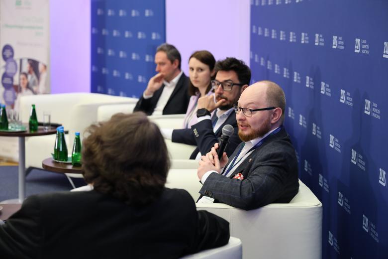 Pięć osób siedzi na fotelach, jedna z nich trzyma w ręku mikrofon i mówi, pozostałe słuchają 