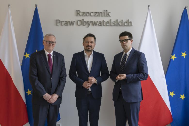 Trzy osoby pozują do wspólnego zdjęcia na tle flag Polski i UE oraz napisu "Rzecznik Praw Obywatelskich" 