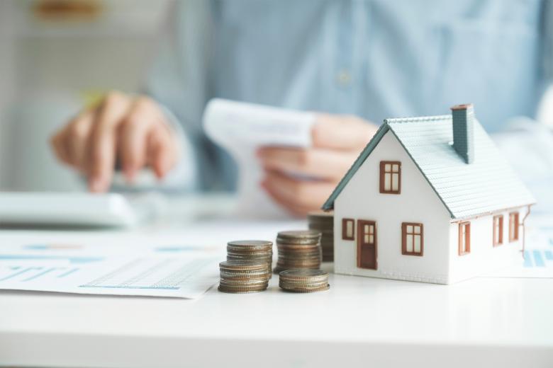 Makieta domu jednorodzinnego i stosy monet leżą na biurku. Na dalszym planie widać osobę czytającą wydruk i korzystającą z kalkulatora.