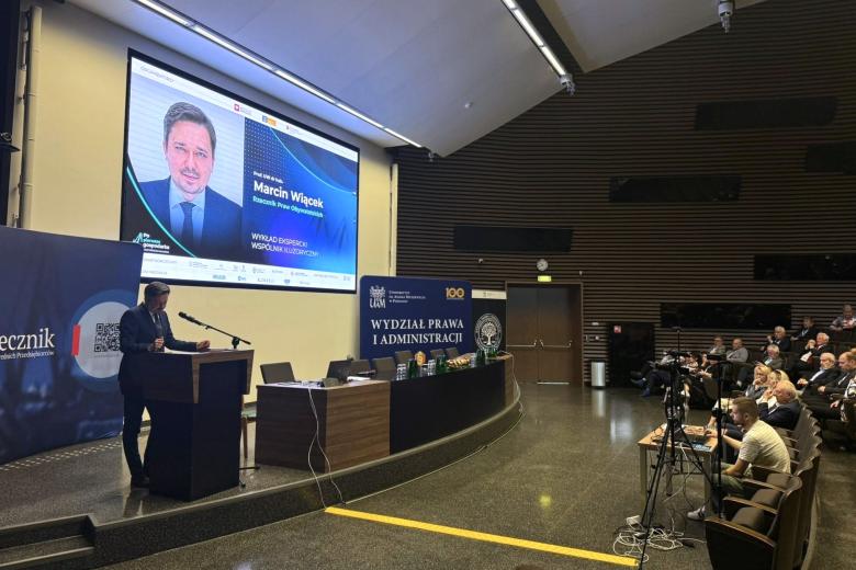 Rzecznik przemawia za mównicą w auli wykładowej, po prawej widać słuchających ludzi, w tle duży ekran z wyświetlonym tematem wystąpienia