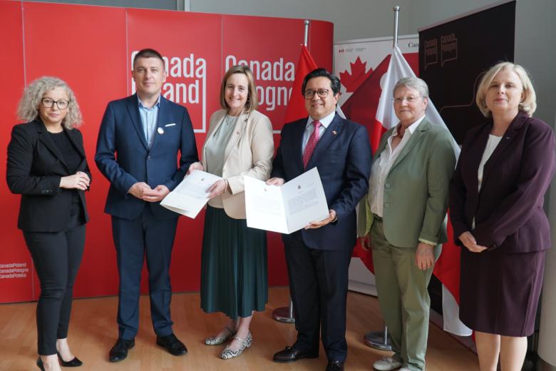 6 osób pozuje do zdjęcia, 2 osoby prezentują list, w tle czerwona ścianka oraz flagi Kanady i Polski