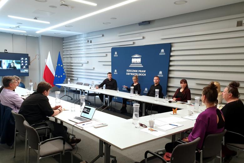 Kilkanaście osób siedzi przy stołach ustawionych w podkowę na sali konferencyjnej, z tyłu wyświetlacz z ekranem programu do komunikowania się na odległość