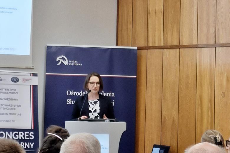 Ewa Dawidziuk stoi przy podium i wypowiada się w stronę słuchającej jej publiczności.