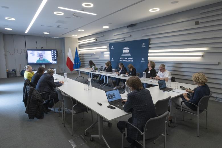 Kilkanaście osób siedzi przy stołach ustawionych w podkowę, na dużym telewizorze na ścianie widoczni uczestnicy posiedzenia łączący się zdalnie za pomocą programu do komunikowania się na odległość