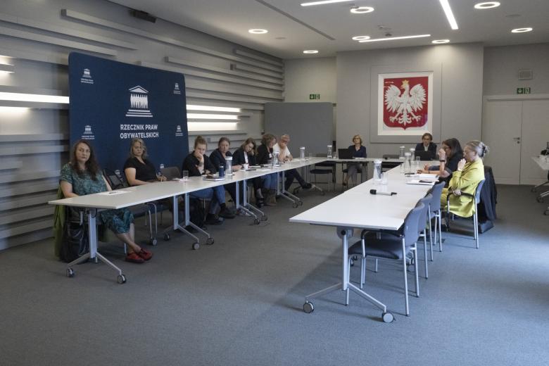 Kilkanaście osób siedzi przy stołach ustawionych w podkowę, za stołami przy ścianie duża plansza z białym napisem Rzecznik Praw Obywatelskich na granatowym tle oraz godło Polski