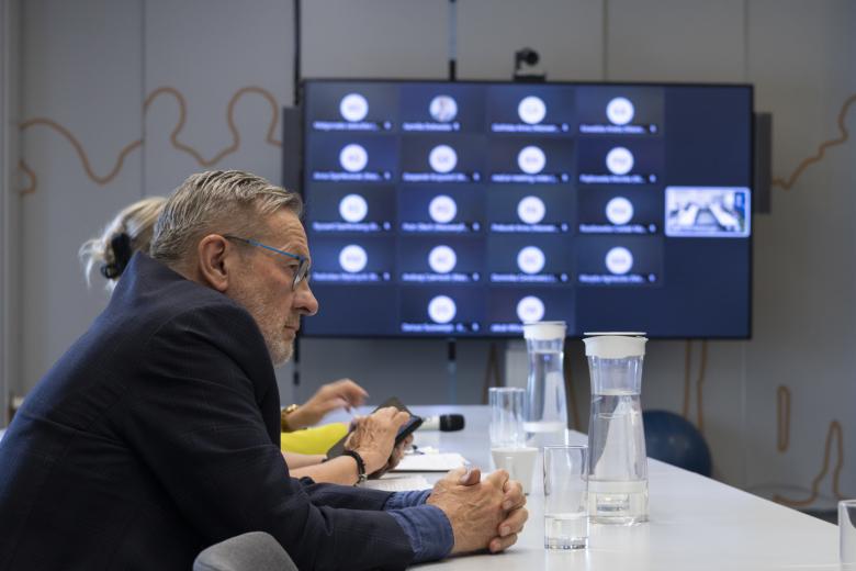 Osoba siedzi przy stole w sali konferencyjnej, a w tle, na dużym telewizorze na ścianie widoczni uczestnicy posiedzenia łączący się zdalnie za pomocą programu do komunikowania się na odległość