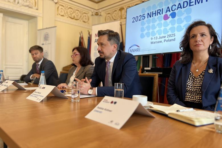 Ujęcie z boku, cztery osoby siedzą przy stole prezydialnym. W tle widać ekran wyświetlający nazwę "2025 NHRI Academy"