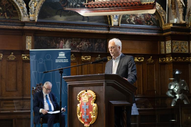 Osoba przemawia za mównicą w zabytkowym, bardzo bogato zdobionym pomieszczeniu w Dworze Artusa w Gdańsku