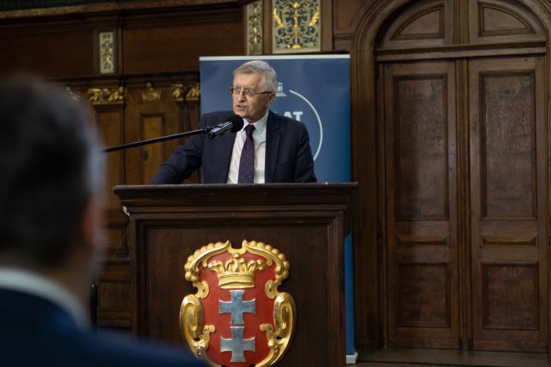 Osoba przemawia za bogato zdobiona mównicą z herbem Gdańska - a czerwonym polu złota korona otwarta i dwa równoramienne srebrne krzyże w słup