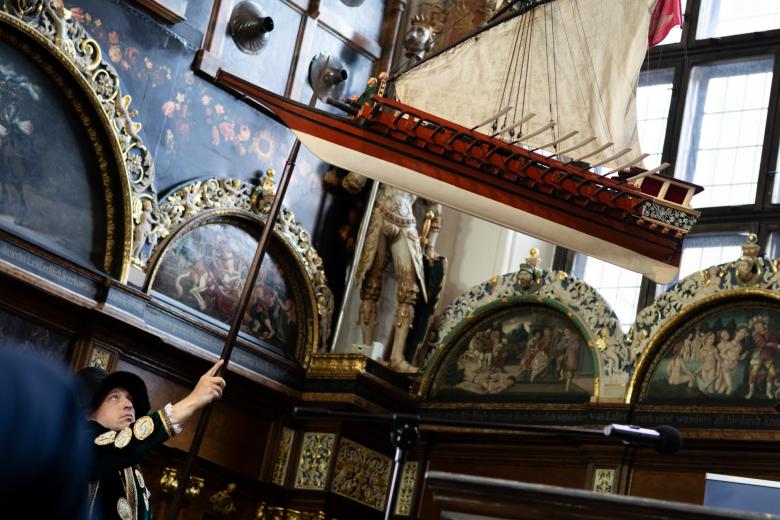 Osoba w historycznym, średniowiecznym stroju podpala lont armaty w modelu galery faluki podwieszonej w sali w zabytkowej, bogato zdobionej sali w Dworze Artusa