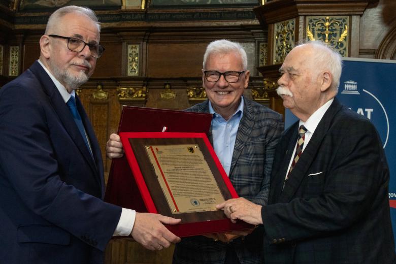 Trzy osoby pozujące do wspólnego zdjęcia trzymające grawer okolicznościowy