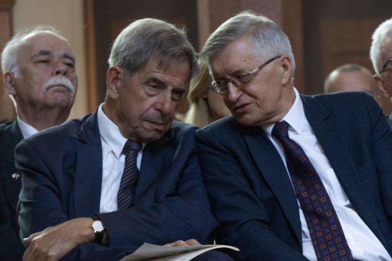 Dwie osoby słuchają wystąpienia siedząc na krzesłach w bogato zdobionej, zabytkowej sali