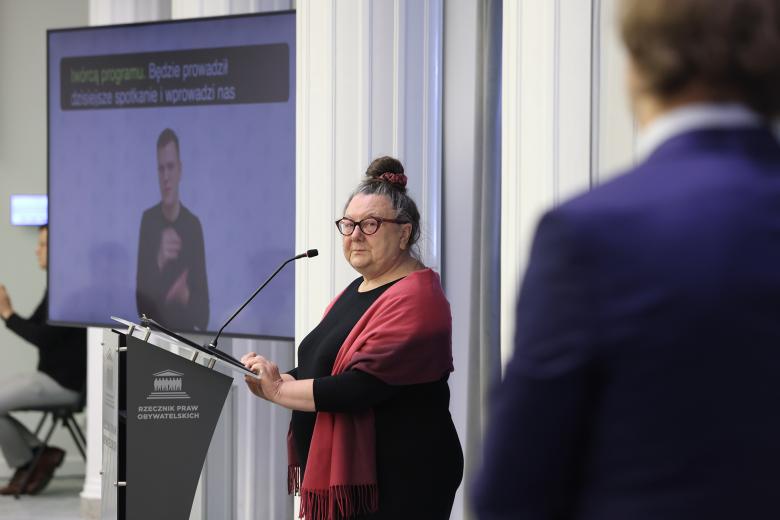 Barbara Imiołczyk wypowiada się stojąc przy mównicy. W tle widać monitor wyświetlający tłumacza języka migowego i napisy.