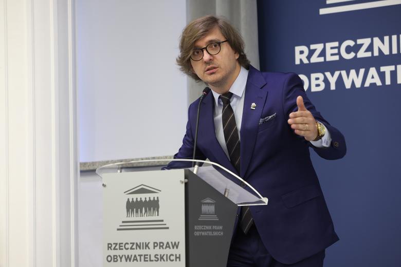 Piotr Zakrzewski stoi przy mównicy i wypowiada się gestykulując