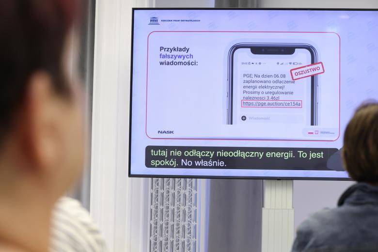 Ekran wyświetlający prezentację, na której widać telefon wyświetlający wiadomość tekstową. Część wiadomości z łączem internetowym jest zakreślona na czerwono z podpisem "oszustwo"