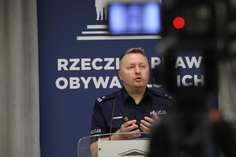 podinsp. Jacek Rybicki wypowiada się stojąc przy mównicy. Na pierwszym planie widać rozmyty zarys nagrywającej go kamery