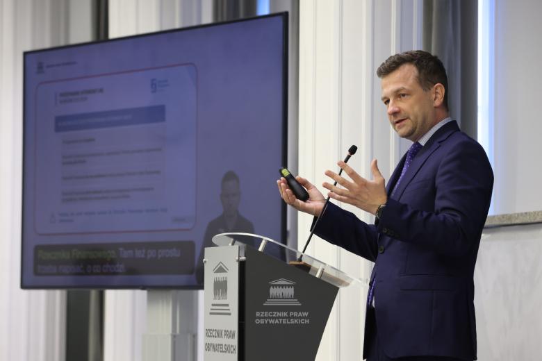 Dawid Rapkiewicz stoi przy mównicy i wypowiada się, gestykulując. W tle widać ekran wyświetlający prezentację, tłumacza języka migowego i napisy