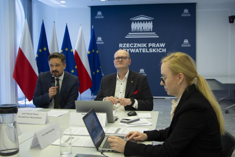 trzy osoby siedzą w sali jedna przemawia na siedząco 