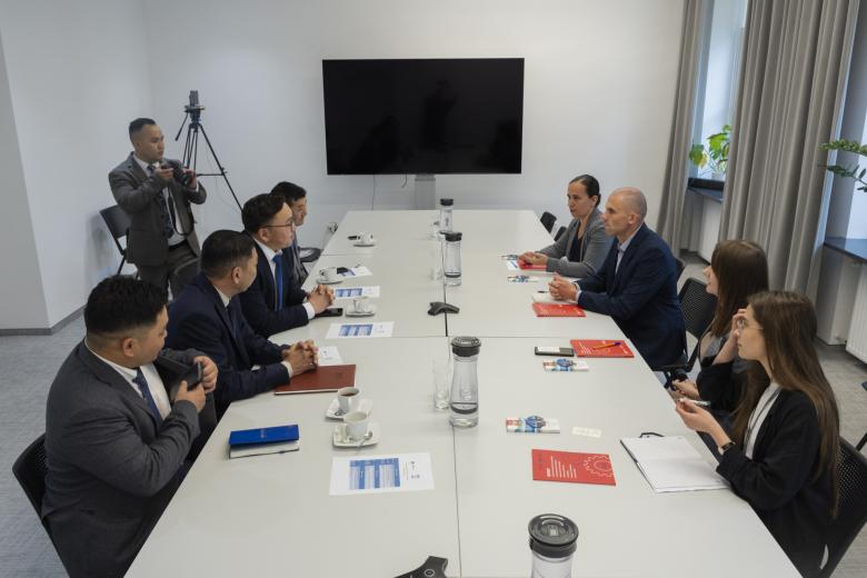 Osiem osób rozmawia siedząc przy stole konferencyjnym. Obok stołu stoi osoba z aparatem fotograficznym w dłoniach