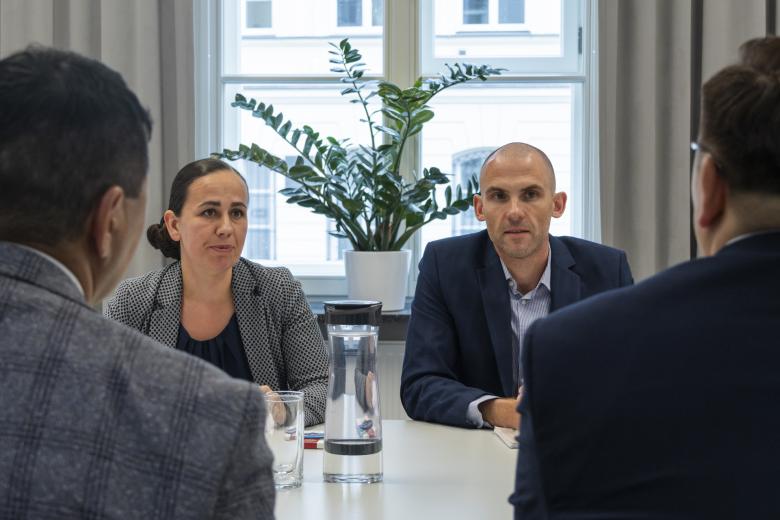 Cztery osoby rozmawiają przy stole konferencyjnym. Widoczne są twarze dwóch osób i sylwetki ich rozmówców 