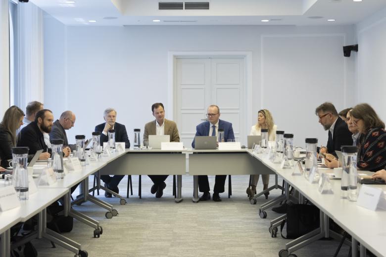 Kilkanaście osób siedzi przy stołach konferencyjnych ustawionych w kształt półkola.