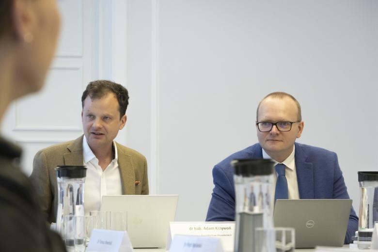 Adam Krzywoń i Piotr Mierzejewski siedzą przy stole konferencyjnym. Adam Krzywoń wypowiada się.