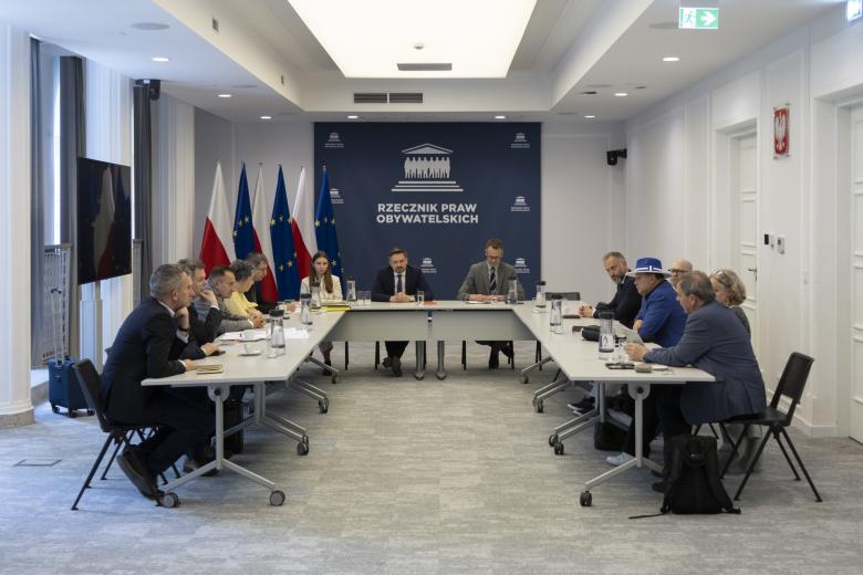 Kilkanaście osób siedzi przy stołach ustawionych w podkowę, za stołami w rogu rząd flag Polski i UE i na środku duża granatowa plansza z białym napisem Rzecznik Praw Obywatelskich