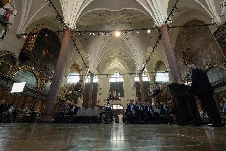 Widok ogólny sali, w której odbywa się konferencja. Przy mównicy stoi wypowiadająca się osoba, przed nią na rzędach foteli siedzą goście. W górze zdjęcia widać gotyckie sklepienie hali.