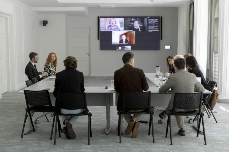 Kilka osób siedzi za stołami ustawionymi w podkowę w sali konferencyjnej