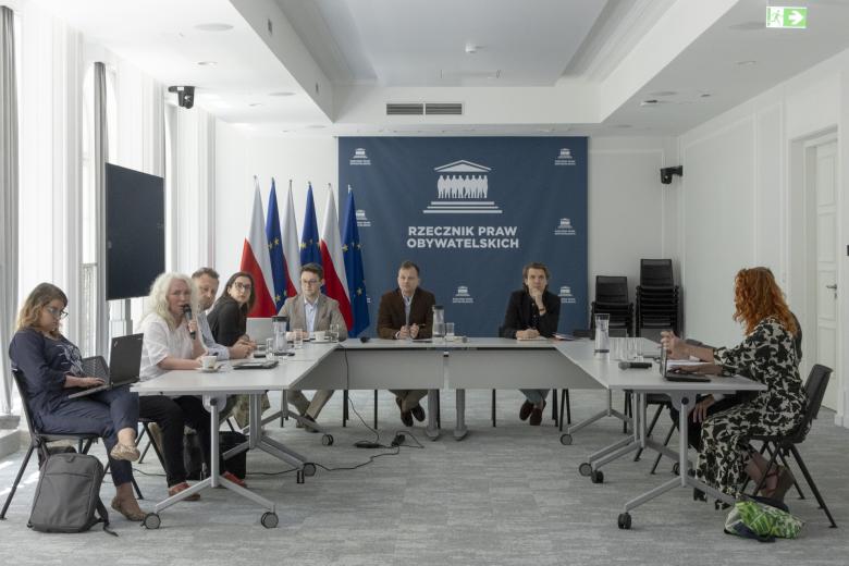 Kilka osób siedzi za stołami ustawionymi w podkowę w sali konferencyjnej