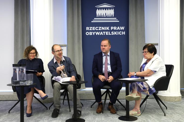 Cztery osoby siedzą na krzesłach przy małych stolikach, jedna z nich mówi do mikrofonu