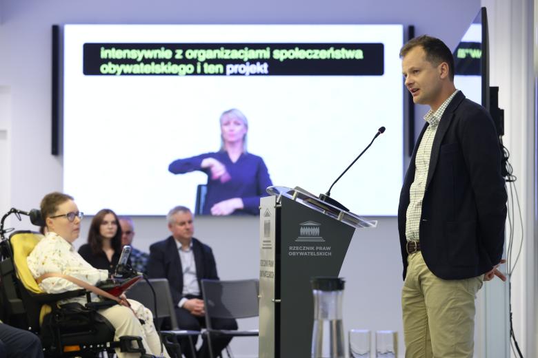 Adam Krzywoń wypowiada się do mikrofonu stojąc przy mównicy. Przed nim siedzą uczestnicy konferencji. W tle widać duży ekran, na którym wyświetlają się napisy oraz tłumacz PJM