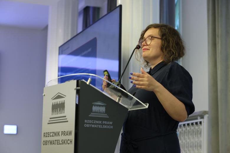 Magdalena Kocejko wypowiada się stojąc przy mównicy i gestykulując