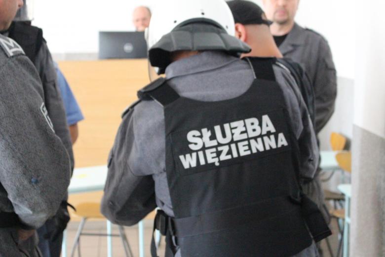 osoba tyłem w kasku i kamizelce z napisem Służba Więzienna obok inne osoby 
